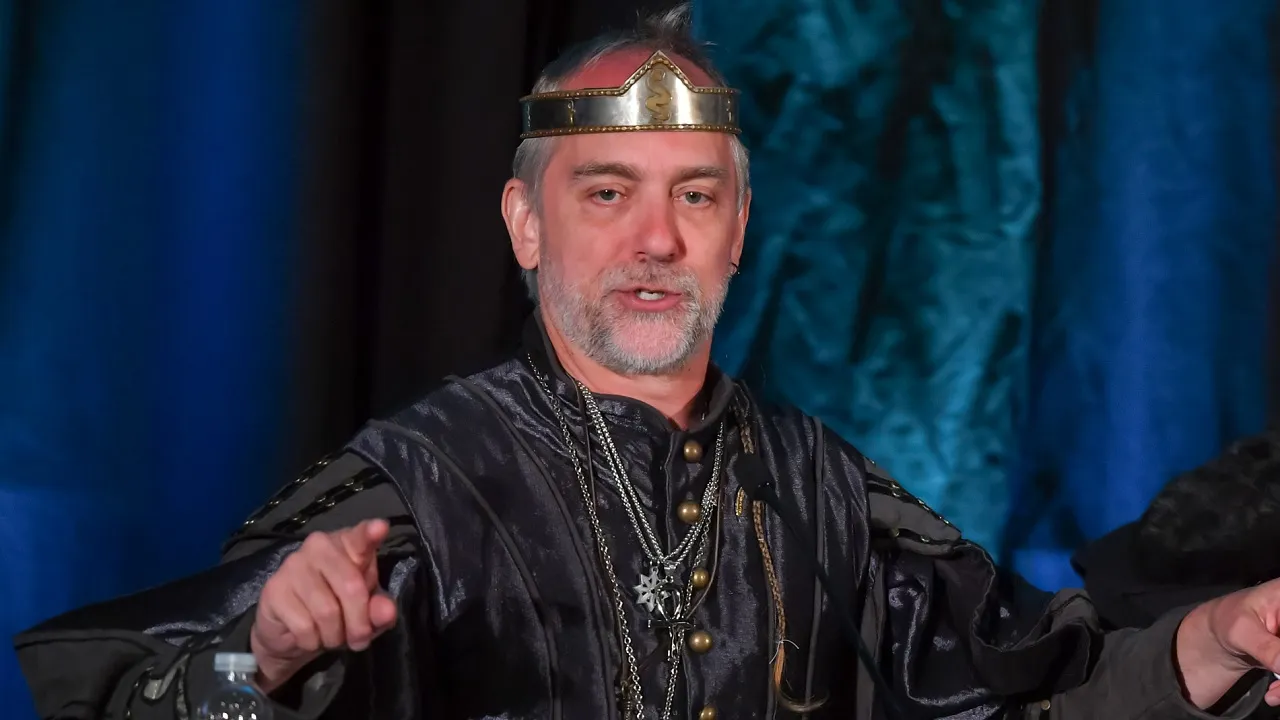 Richard Garriott in a black medieval-style tunic and a metal circlet, speaking and pointing on stage in front of blue and teal curtains